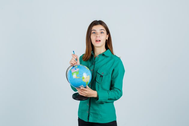 指向年轻的女士拿着地球仪 指着它在衬衫上 看起来困惑 前视图拥抱肖像时尚