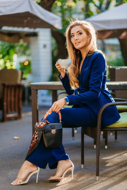 鞋子穿着蓝色优雅套装的迷人时尚女士坐在咖啡馆的桌子旁喝着一杯卡布奇诺秋天西装街头风格
