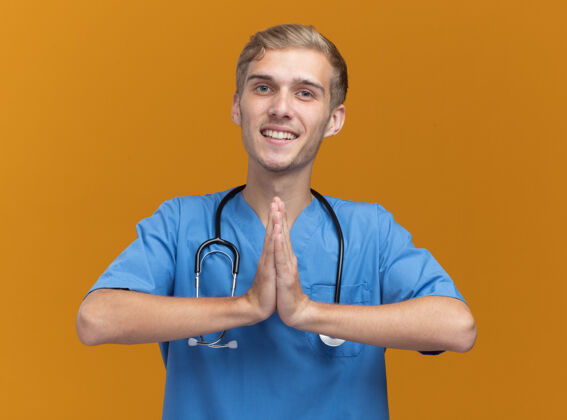 展示微笑的年轻男医生穿着医生制服 听诊器显示祈祷手势 隔离在橙色墙上佩戴医生男性