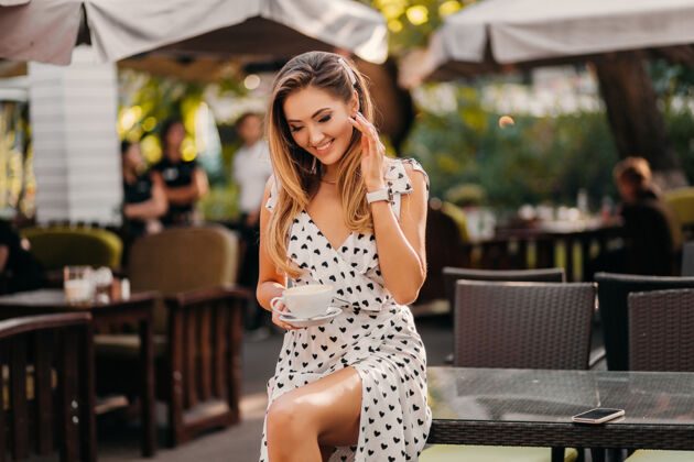 灯光美丽的女人带着坦率可爱的微笑坐在咖啡馆里 手里拿着一杯卡布奇诺 穿着印花时尚的白色连衣裙服饰桌子年轻