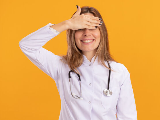 穿面带微笑的年轻女医生穿着医用长袍 听诊器遮住眼睛 手被隔离在黄色的墙上医生听诊器年轻