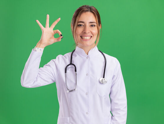 听诊器微笑的年轻女医生穿着医用长袍 听诊器显示出良好的姿态隔离在绿色的墙上医生表演长袍