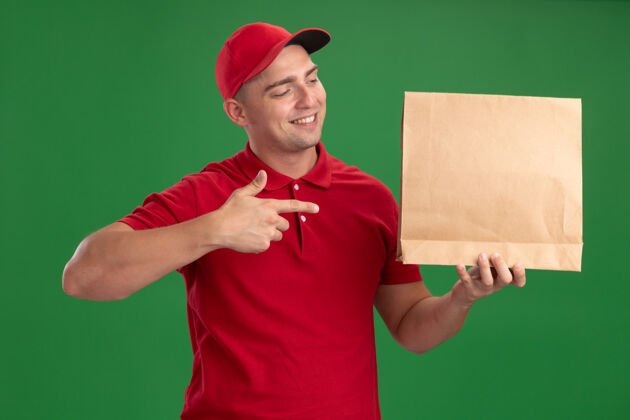微笑面带微笑的年轻送货员穿着制服 戴着帽子 指着隔离在绿色墙上的纸制食品包拿制服包装