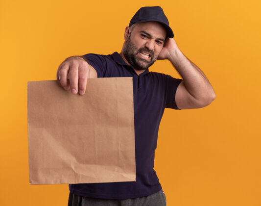 纸张关心的中年送货员穿着制服 戴着帽子 看着纸食品包装把手放在头上隔离在黄色的墙上中年制服送货