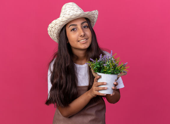 立场戴着围裙和夏帽的快乐的年轻园丁女孩 手拿盆栽植物 脸上带着微笑站在粉红色的墙上花园围裙锅