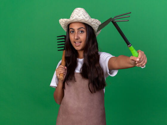 年轻快乐的年轻园丁女孩 围裙 戴着夏帽 手里拿着小耙子 面带微笑地站在绿色的墙上