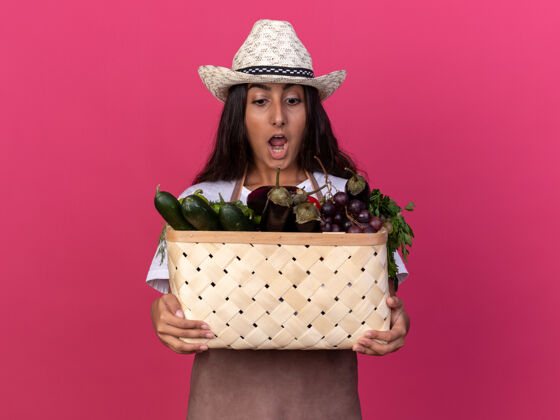 立场年轻的园丁女孩围着围裙 戴着夏帽 手里拿着装满蔬菜的板条箱 站在粉色的墙上惊讶地看着它们围裙惊人蔬菜