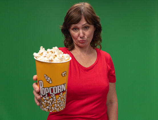 中年一位身穿红色t恤的中年妇女站在绿色的墙上 手里拿着一桶爆米花 表情悲伤年龄表情桶