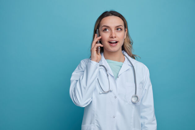 年轻兴奋的年轻女医生穿着医用长袍 脖子上戴着听诊器 抬头看电话长袍周围电话