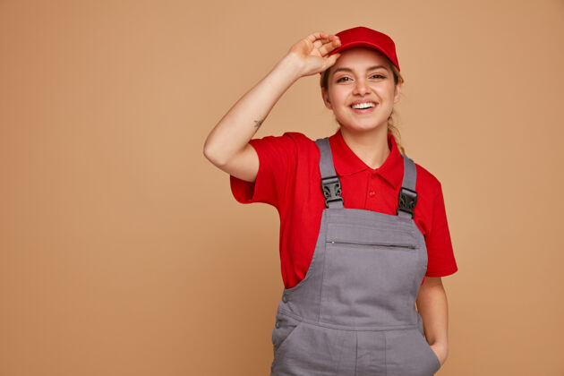 复制快乐的年轻女建筑工人戴着帽子和制服 手放在口袋里抓着帽子工人口袋建筑