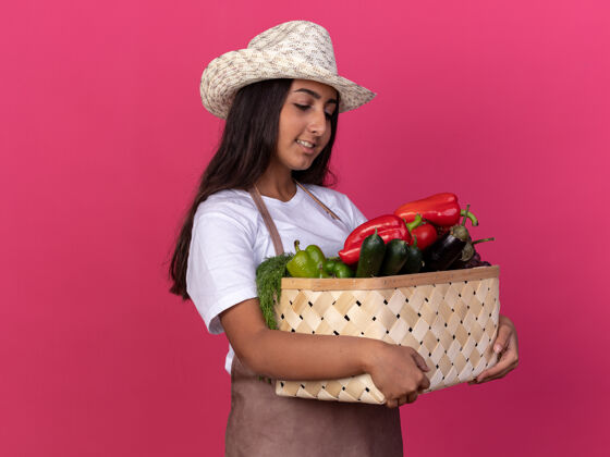 花园年轻的园丁女孩围着围裙 戴着夏天的帽子 手里拿着装满蔬菜的箱子 站在粉色的墙上 看上去自信地微笑着满的架子蔬菜
