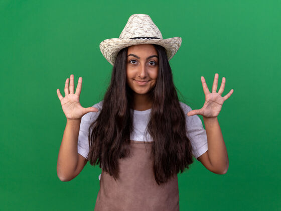 显示穿着围裙 戴着夏帽的年轻园丁女孩微笑着 张开手掌站在绿色的墙上棕榈花园欢呼