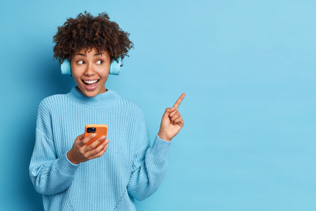 音乐帅气的非裔美国女人的横拍感觉很开心手持现代智能手机戴着立体声耳机在蓝色背景的空白处指指点点休闲概念指向高兴空白