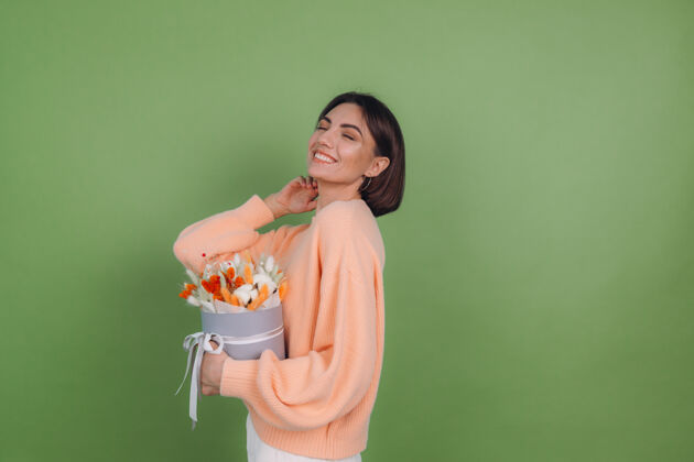 欢乐年轻女子穿着休闲桃色毛衣隔离在绿橄榄墙上手持橙白色花盒组成的棉花 吉普赛拉小麦和拉古鲁斯作为礼物开心惊喜女人黑麦干花