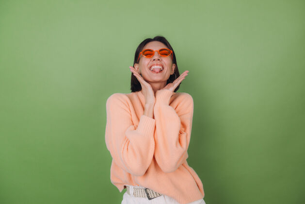 模特年轻时尚的女士穿着休闲的桃色毛衣 戴着橙色的眼镜 隔着绿橄榄色的墙壁 开心的 积极的笑着 围着复制空间转魅力积极黑发