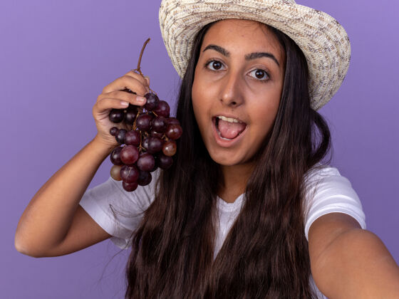 幼崽穿着围裙 戴着夏帽 手里拿着一束葡萄的年轻园丁姑娘站在紫色的墙上 脸上洋溢着幸福的笑容串架子花园