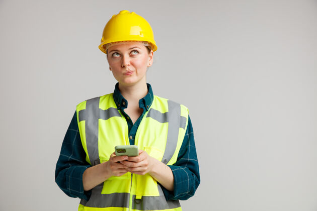电话体贴的年轻女建筑工人戴着安全帽和安全背心拿着手机噘着嘴唇仰望空间钱包复制