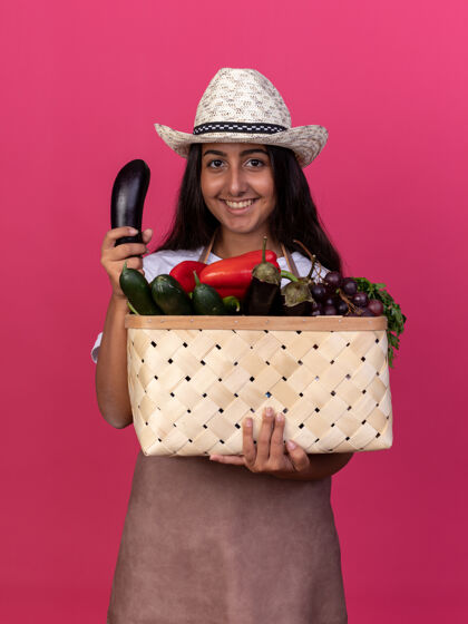 茄子快乐的年轻园丁女孩 围着围裙 戴着夏帽 手里拿着装满蔬菜和新鲜茄子的箱子 脸上带着微笑站在粉红色的墙上花园立场围裙