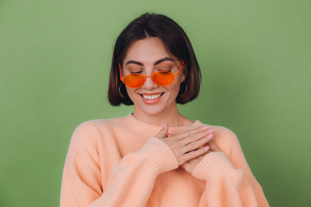 温柔年轻时尚的女士穿着休闲桃色毛衣 戴着橘色眼镜 隔着绿橄榄色的墙正面双手合十放在胸前 心复制空间人成人微笑