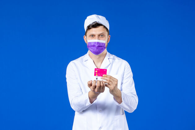 男医生前视图中的男医生穿着医疗服和紫色面罩持有注射和信用卡在蓝色信用卡药品钱