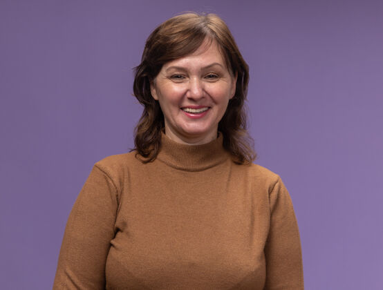 宽的快乐的中年妇女穿着棕色高领毛衣站在紫色的墙上 面带微笑高领年龄中等