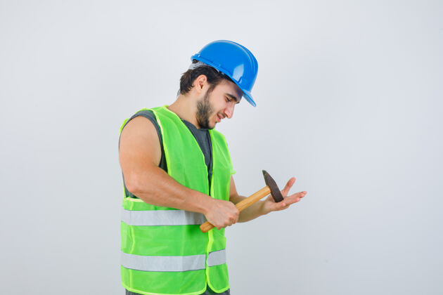 男孩年轻的建筑工人穿着工作服在手掌上敲打锤子 看上去很高兴 正面视图前面模特手掌