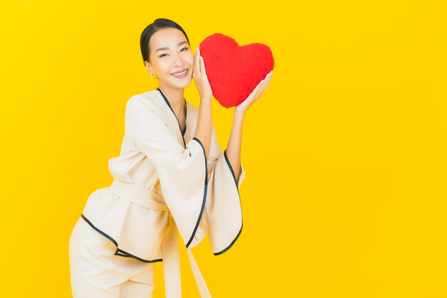 浪漫黄色墙壁上有一个心形枕头的亚洲商界年轻美女的画像爱快乐模特