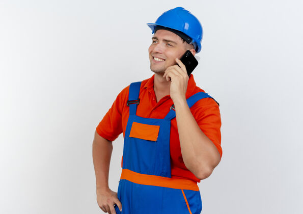 手看着面带微笑 身穿制服 头戴安全帽的年轻男性建筑工人一边讲电话 一边把手放在屁股上男性男人头盔
