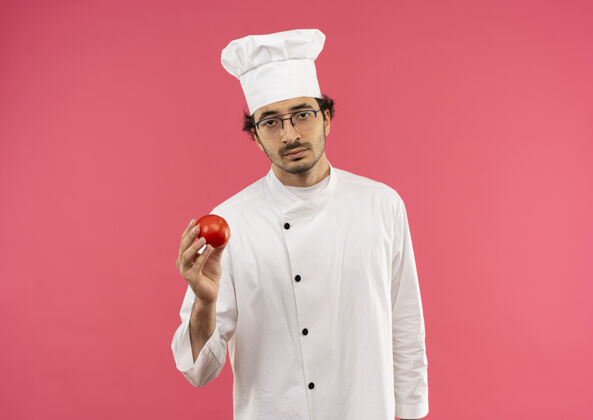 粉红悲伤的年轻男厨师穿着厨师制服 戴着眼镜拿着西红柿烹饪男年轻