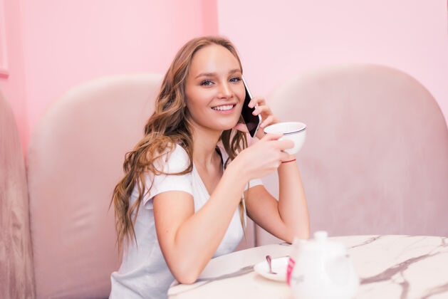 坐着一位身着灰色礼服的年轻女商人坐在咖啡馆的桌子旁 一边用手机聊天 一边在笔记本上做笔记女性笔记本电脑教育