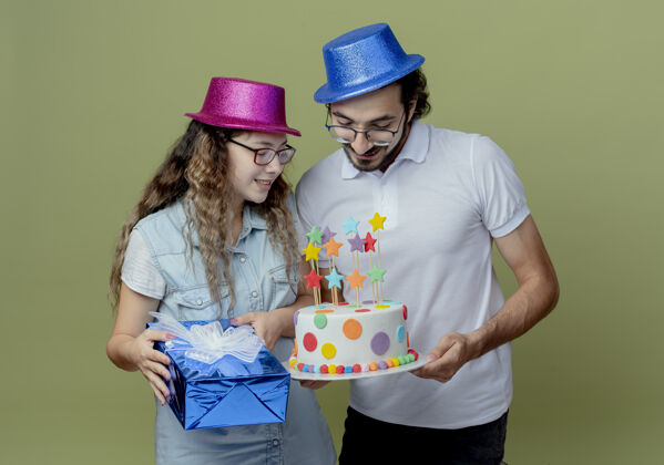 戴着高兴的年轻夫妇戴着粉红色和蓝色的帽子 女孩拿着礼品盒 男孩抱着和女孩一起看生日蛋糕帽子蛋糕绿色