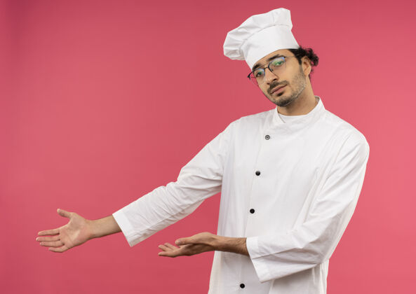 粉红年轻的男厨师穿着厨师制服 戴着眼镜点着手边点穿烹饪