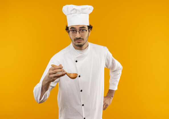 眼镜年轻的男厨师穿着厨师制服 戴着眼镜 手举勺子放在臀部穿着手持厨师