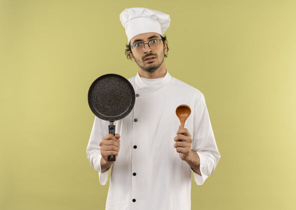 平底锅年轻的男厨师穿着厨师制服 戴着眼镜 手里拿着煎锅和勺子薯条男性持有