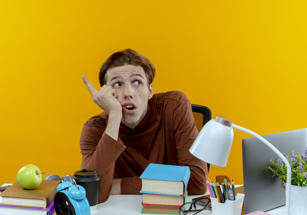 书桌看着边思考的年轻学生男孩坐在书桌旁 手里拿着学校的工具指着旁边黄色思考年轻