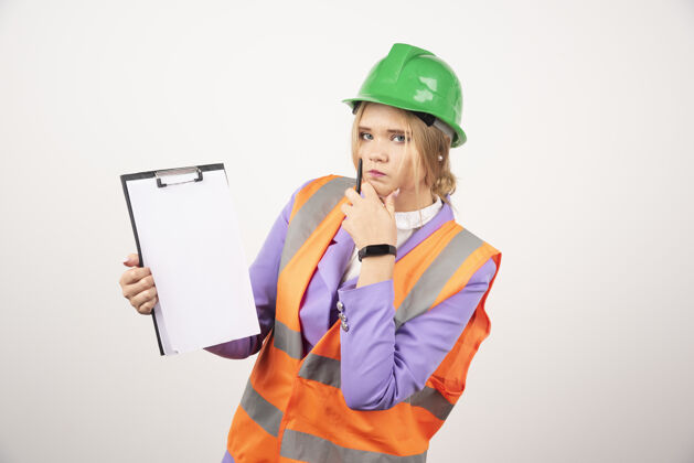 工作年轻的女承包商 戴着绿色头盔 白墙上有剪贴板工程师女性女孩