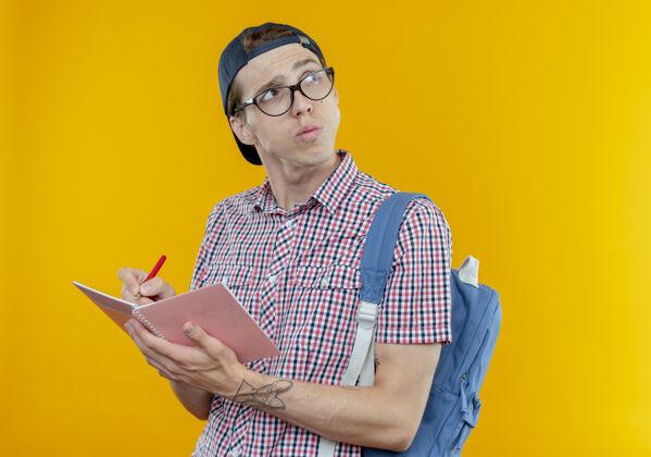 青少年看着边思考的年轻学生男孩 背着背包 戴着眼镜 戴着帽子 拿着笔记本和钢笔男人男性穿
