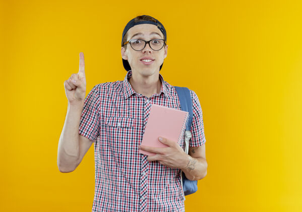 笔记本印象深刻的年轻学生男孩背着背包 戴着眼镜和帽子拿着笔记本 指着上面极客帽子点