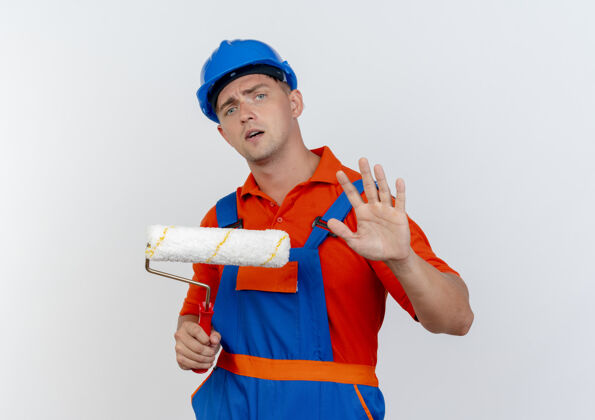 关心关心的年轻男性建筑工人身穿制服 戴安全帽 手持油漆辊 并显示停止手势年轻人安全滚轴