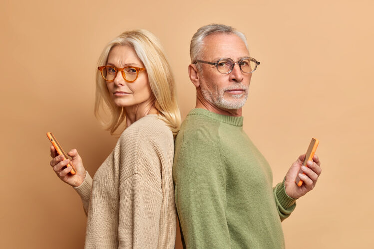 在一起严肃的老太太和她的丈夫拿着现代化的小玩意看媒体闲暇时间在互联网上互相忽略站在后面戴着眼镜毛衣隔着米色的墙老人男家庭