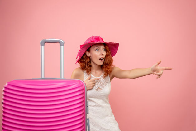 美丽年轻的女性游客与粉红色的帽子和粉红色的墙壁上的包的正面视图帽子快乐微笑