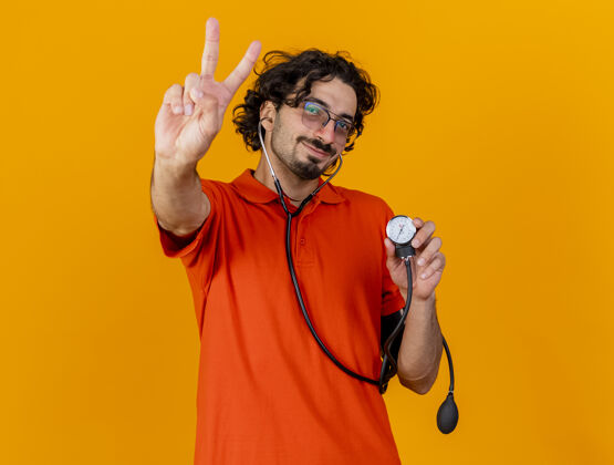橙色高兴的年轻白种人病人戴着眼镜和听诊器拿着血压计做和平的标志隔离在橙色墙上的复制空间请戴上和平疾病