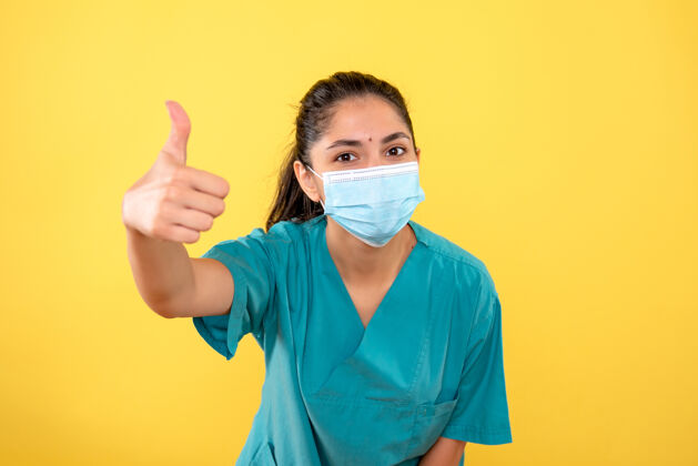 医学戴着医用面罩的年轻女性正面图 黄色墙上有竖起拇指的标志装置拇指肖像