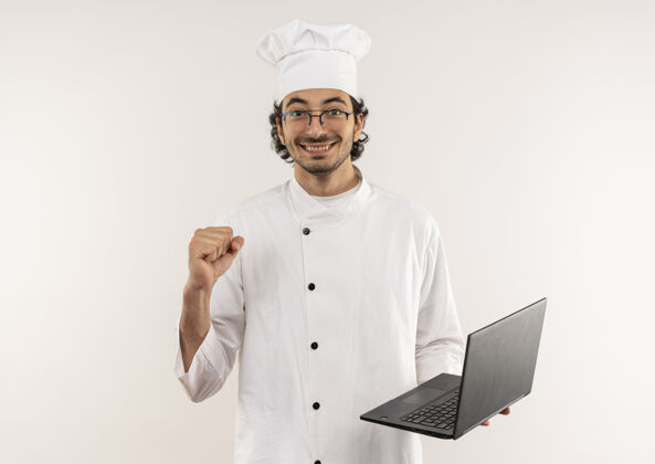手持面带微笑的年轻男厨师穿着厨师制服 戴着眼镜 拿着笔记本电脑 在白色的墙上显示“是”的手势手势笔记本电脑烹饪