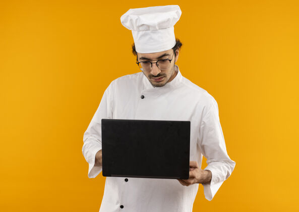 持有想着年轻的男厨师穿着厨师制服戴着眼镜拿着笔记本电脑看着隔离在黄色墙上烹饪厨师眼镜