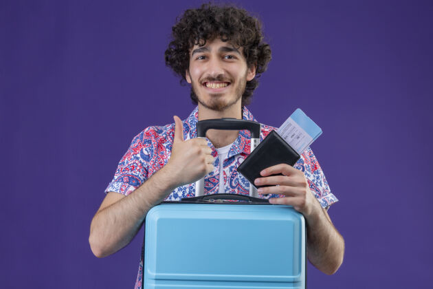 帅气微笑着的年轻英俊的卷发旅行者 手里拿着机票和钱包 手提箱在孤立的紫色墙上竖起大拇指钱包年轻人抱着