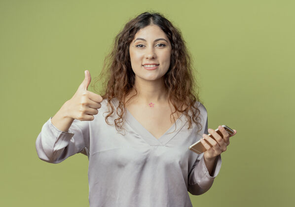拇指微笑着的年轻漂亮的女办公室工作人员举着电话 她的大拇指孤立地放在橄榄绿的墙上拿着微笑电话