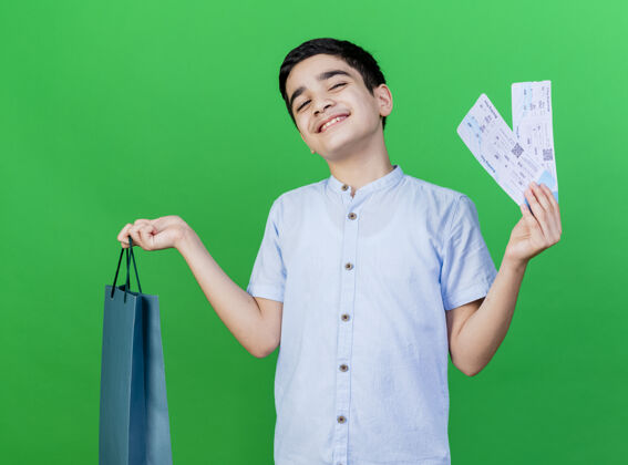 年轻人高兴的年轻白人男孩拿着购物袋和机票隔离在绿墙上购物男人男孩