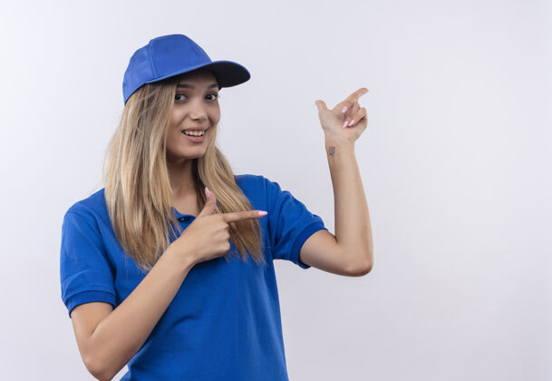 穿面带微笑的年轻送货员身穿蓝色制服 戴着帽子 指着隔离在白色墙上的一面 留着复印空间交货制服女孩