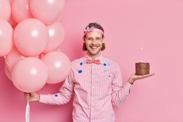 眼镜兴高采烈的留着时髦发型的没刮胡子的家伙准备庆祝生日 手里拿着巧克力蛋糕和氦气球在派对上玩得很开心 收到朋友们的祝贺五彩纸屑飞来飞去单身帅哥蜡烛